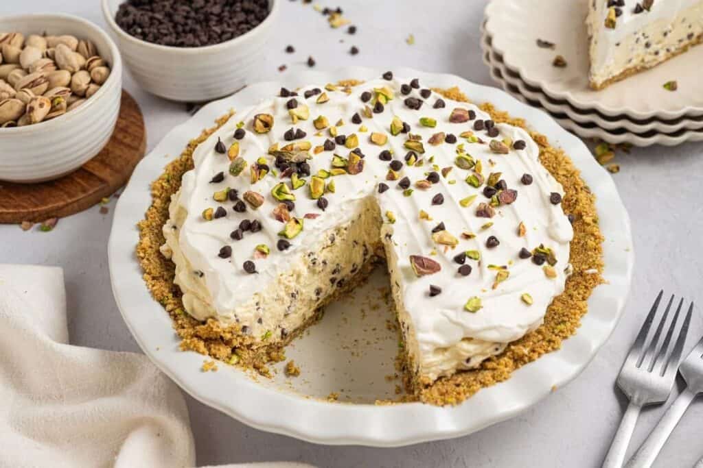 A no bake treat, this cream pie with a slice missing is topped with whipped cream, chopped pistachios, and chocolate chips, and sits in a white dish next to bowls of pistachios and chocolate chips.