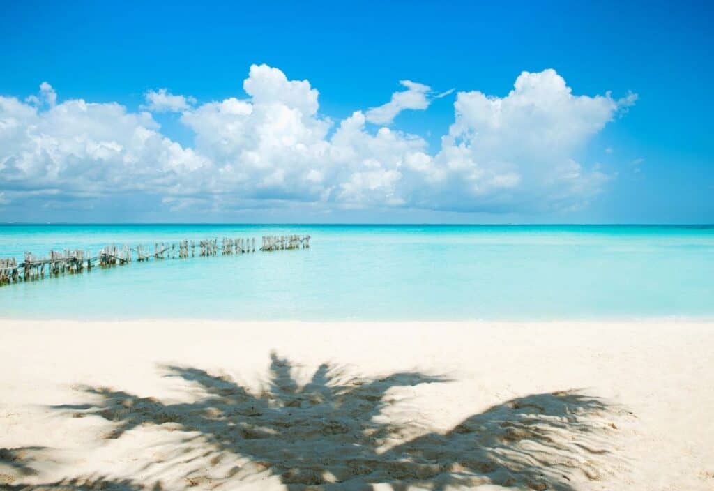 White sand beach with clear turquoise water, a wooden pier to the left, palm tree shadow in the foreground, and blue sky with scattered clouds.