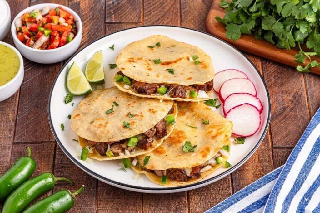 A plate with three quesadillas filled with meat and vegetables, inspired by tasty beef recipes, garnished with chopped cilantro, lime wedges, and sliced radishes, surrounded by salsas and fresh cilantro.