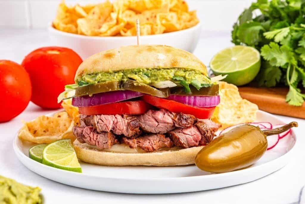 A steak sandwich with sliced tomatoes, red onions, pickles, and guacamole on a bun&mdash;one of our tasty beef recipes&mdash;served with tortilla chips, lime wedges, and a jalape&ntilde;o on a white plate.