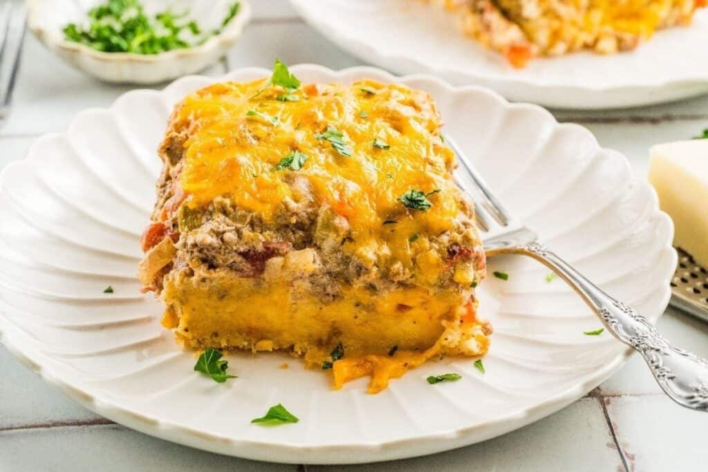 A square slice of cheesy casserole, inspired by tasty beef recipes, is topped with chopped parsley and served on a white plate with a fork.