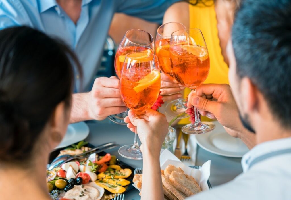 Four people raise glasses of orange-colored drinks in a toast over a table set with food, including bread and salad.