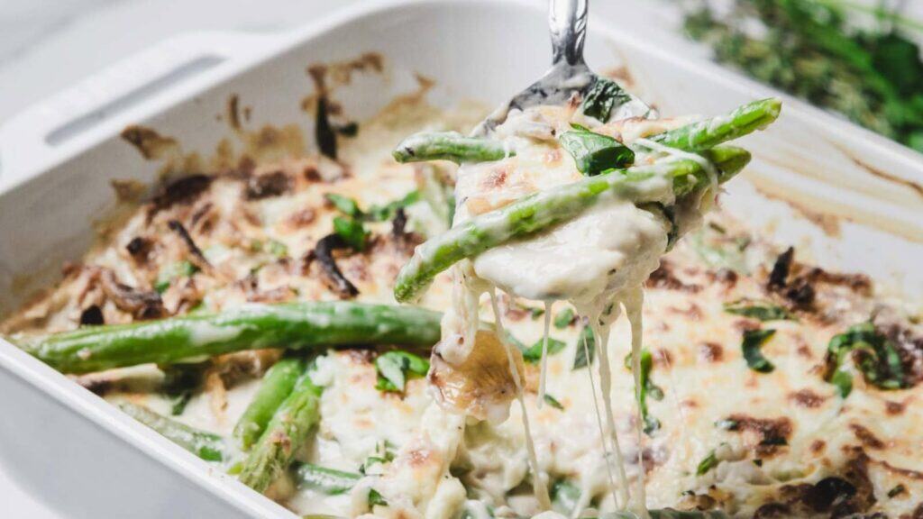 A cheesy green bean casserole with melted cheese on top, served in a white baking dish. A fork holds up a portion with green beans and creamy sauce.