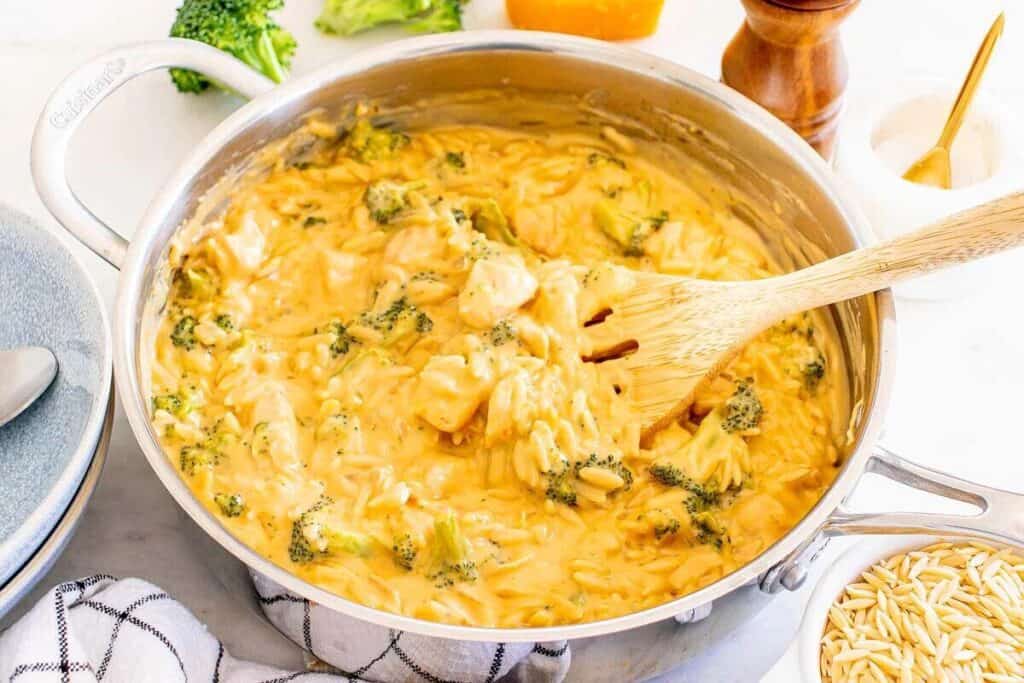 A saucepan filled with creamy orzo pasta, broccoli, and chicken, being stirred with a wooden spoon on a white countertop.