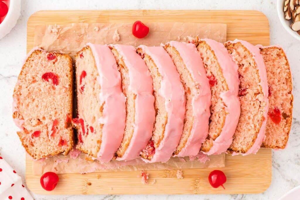 Sliced cherry bread with pink icing is arranged on a wooden cutting board, with whole cherries scattered around&mdash;perfect for those who love sweet breakfast recipes.