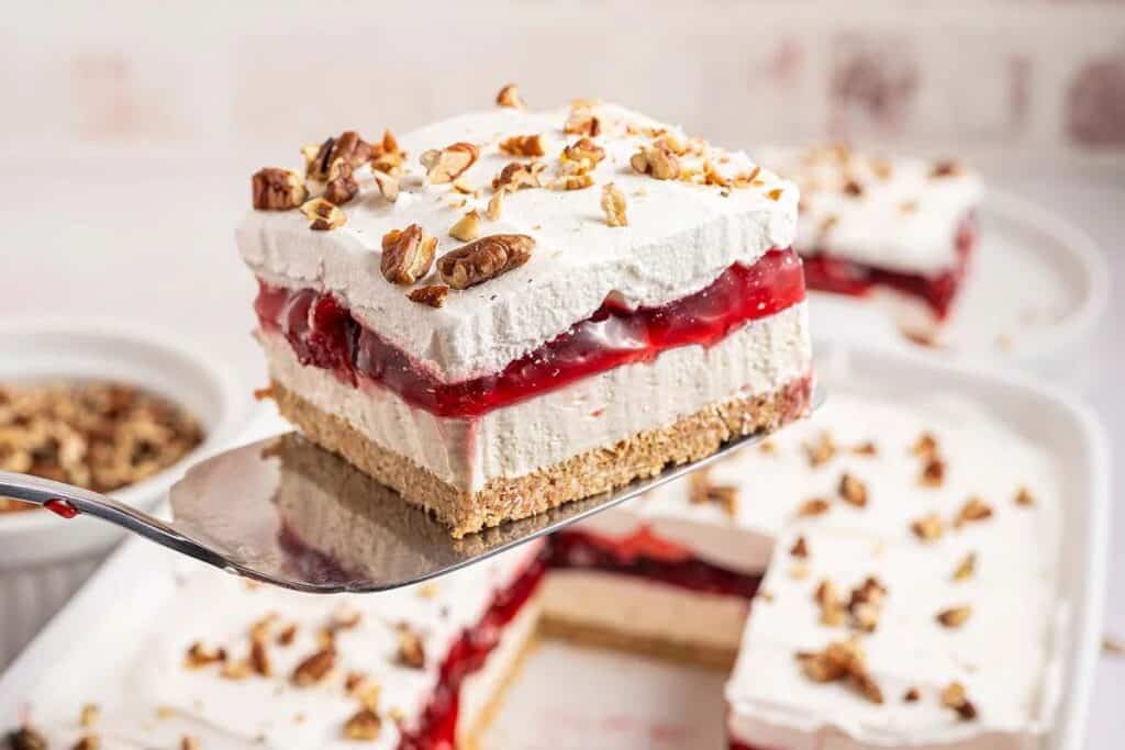 A spatula holds a slice of layered dessert with a graham cracker crust, creamy filling, cherry topping, whipped cream, and chopped pecans&mdash;one of the ultimate no bake treats. Other slices are visible in the background.