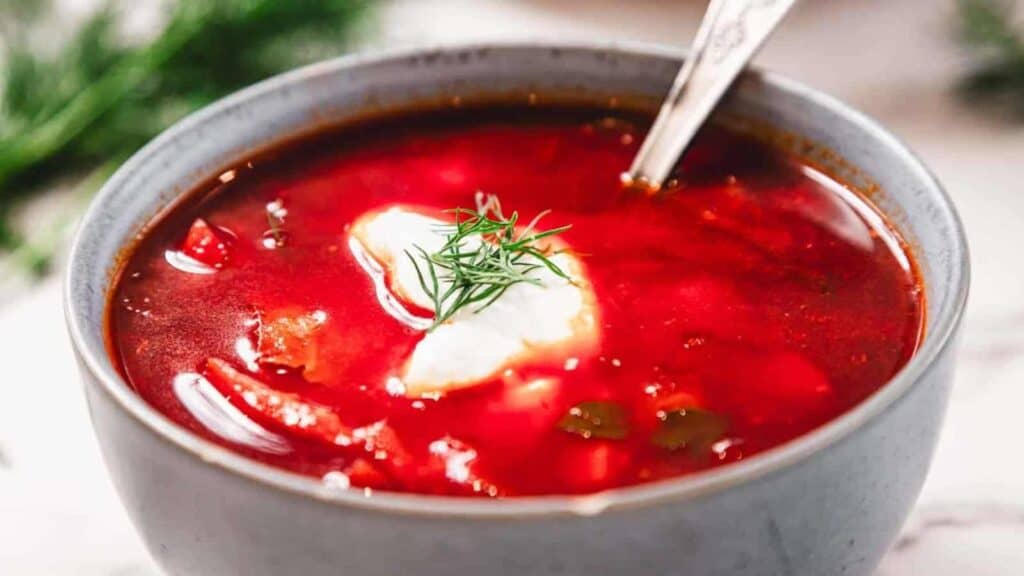 A bowl of red soup, likely borscht, topped with a dollop of sour cream and a sprig of dill, with a spoon in the bowl.