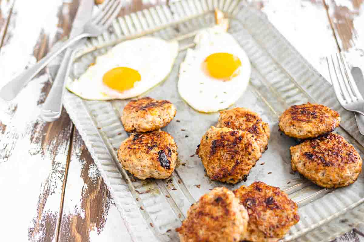 A metal tray with eight browned sausage patties and two sunny-side-up eggs, with forks and knives placed nearby on a rustic wooden table.