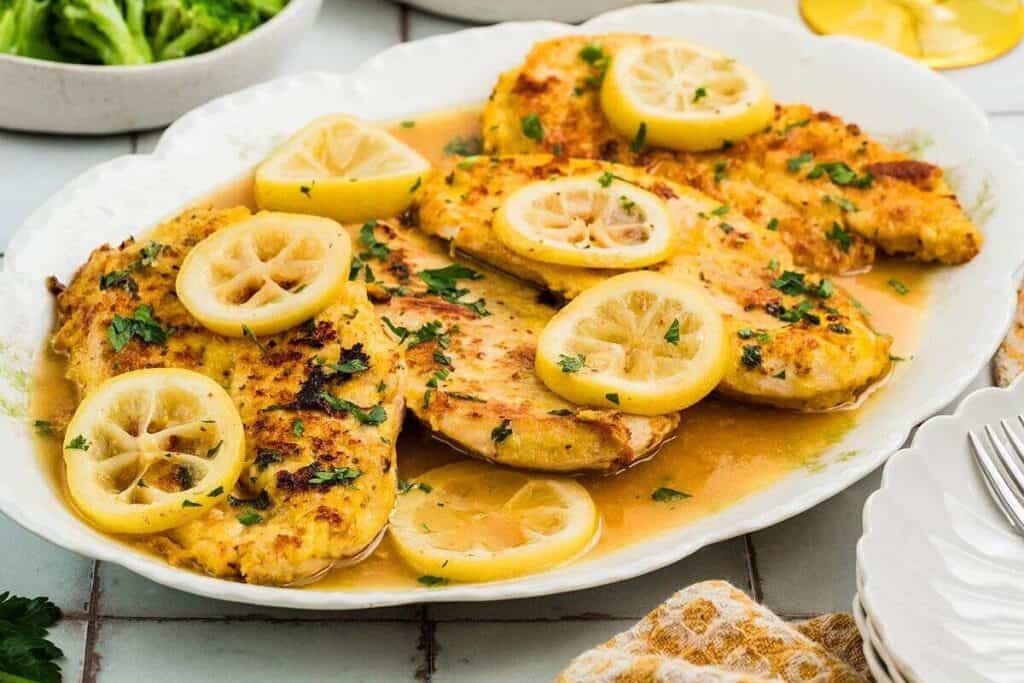 Grilled chicken breasts topped with lemon slices and chopped parsley served on a white platter with a lemon sauce.