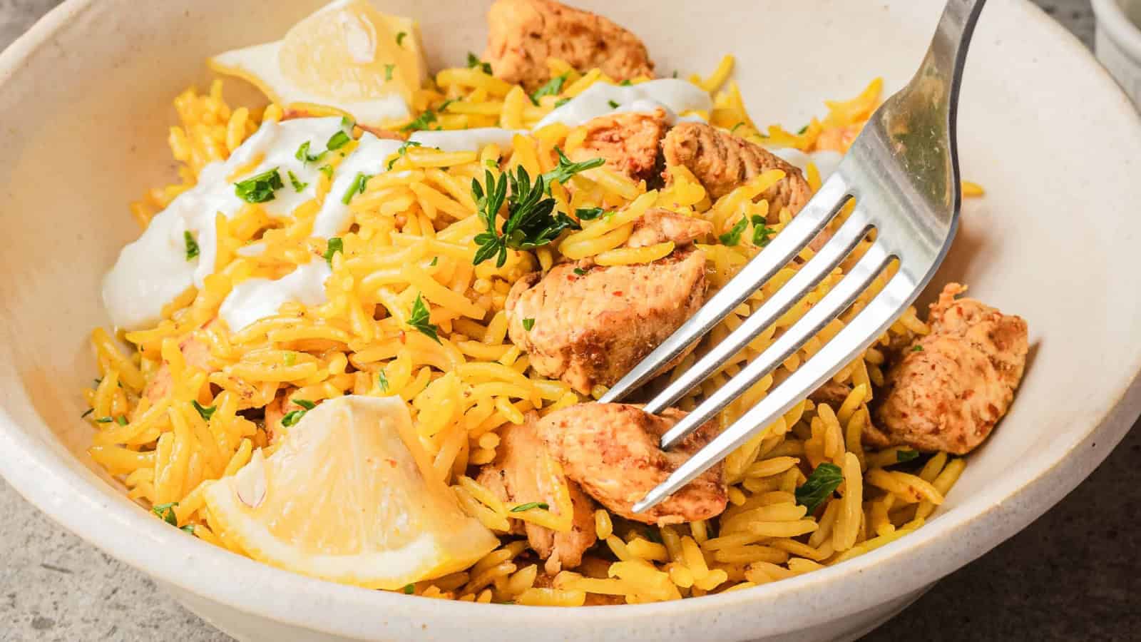 A fork in a bowl of yellow rice with grilled chicken, lemon wedges, herbs, and a dollop of white sauce.