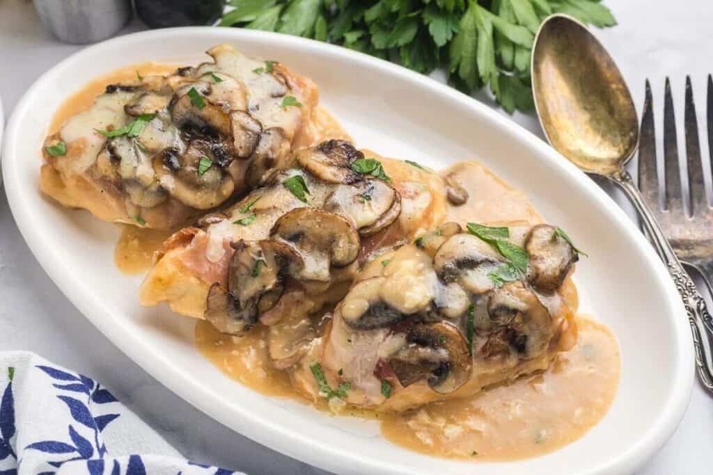 Three pieces of chicken topped with mushrooms and melted cheese are served on a white oval plate with a creamy sauce, garnished with parsley.