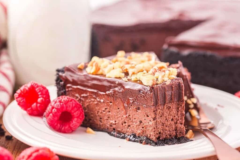 A slice of simple cheesecake, rich with chocolate and topped with chopped nuts, sits on a plate with fresh raspberries, accompanied by a fork and the remaining cake in the background.