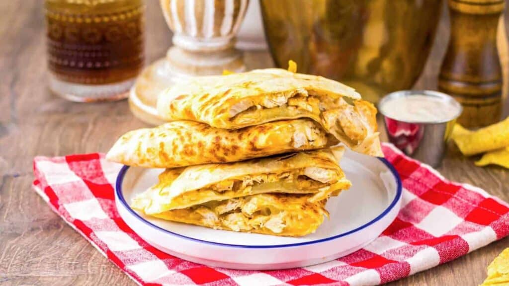 Three pieces of stuffed flatbread with a golden crust are stacked on a white plate atop a red-and-white checkered napkin, with a cup of dipping sauce nearby.