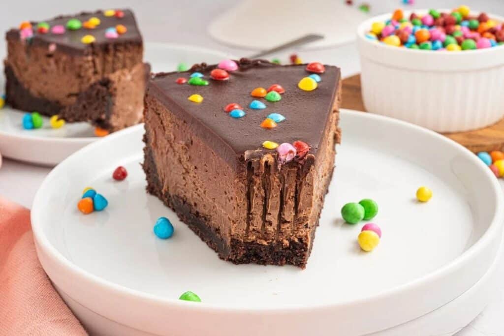 A slice of Simple Cheesecake with a rich chocolate ganache topping and colorful candy pieces sits on a white plate.