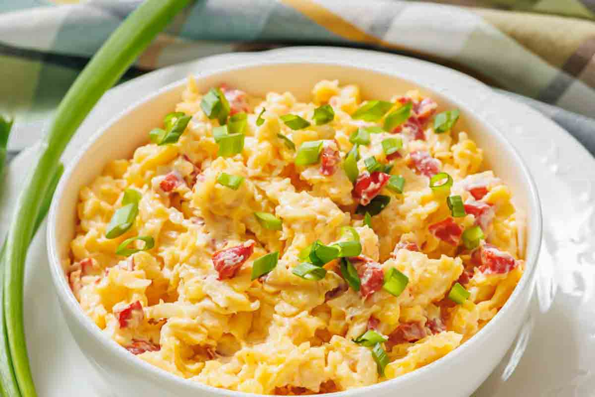 A bowl of pimento cheese spread with diced red bell peppers and topped with chopped green onions, set on a white plate.