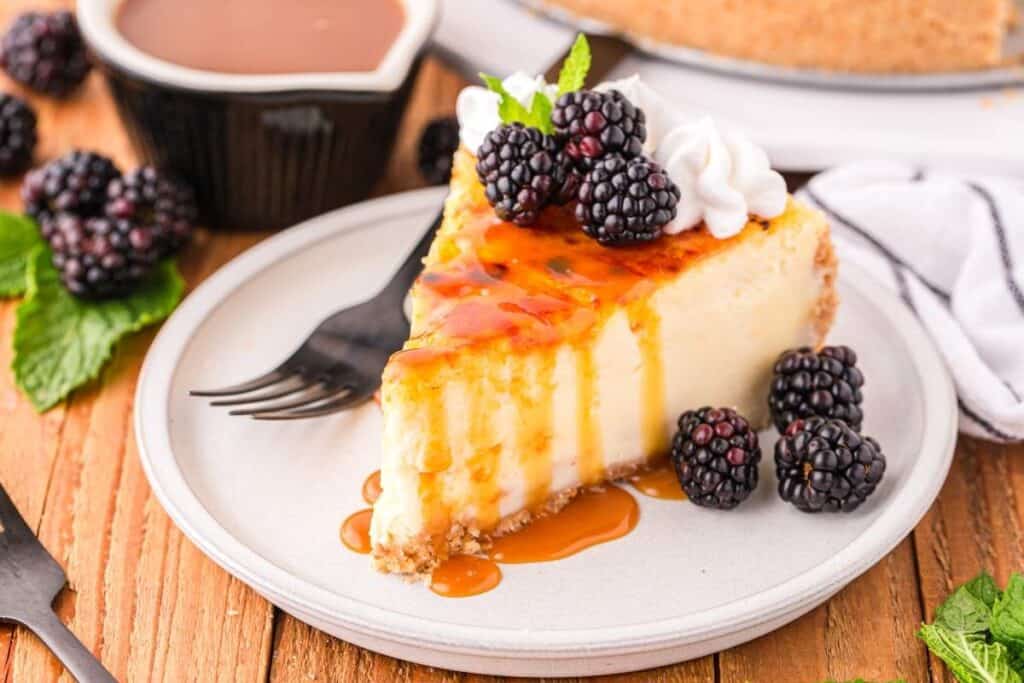 A slice of Simple Cheesecake topped with blackberry, whipped cream, and caramel sauce sits on a plate with a fork and fresh blackberries on the side.