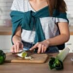A woman slices a leek on a wooden cutting board in a kitchen, surrounded by fresh vegetables like bell pepper, broccoli, and carrots.