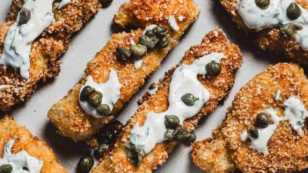 Breaded fish sticks topped with white sauce and capers are arranged on a light-colored surface.