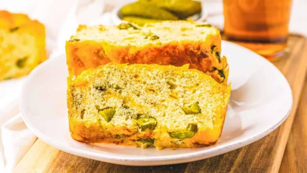 Two thick slices of savory bread with visible pieces of pickles on a white plate, set on a wooden surface.
