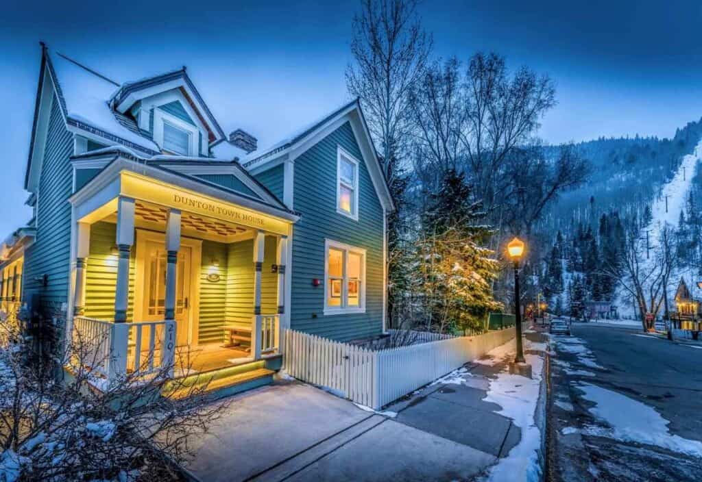 A green Victorian-style house with a lit porch named “Dunton Town House,” surrounded by snow, trees, and a mountain slope at dusk—an inviting retreat that embodies the charm of ultra-boutique hotels.