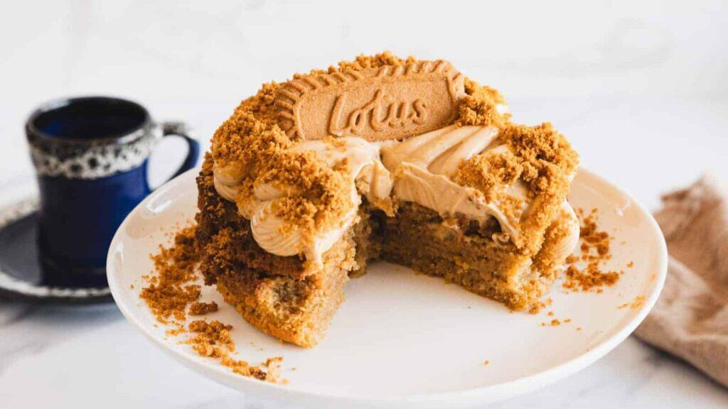 A partially sliced Lotus Biscoff cake with buttercream and crumble topping sits on a white cake stand next to a dark cup of coffee, perfect inspiration for those exploring new air fryer recipes.