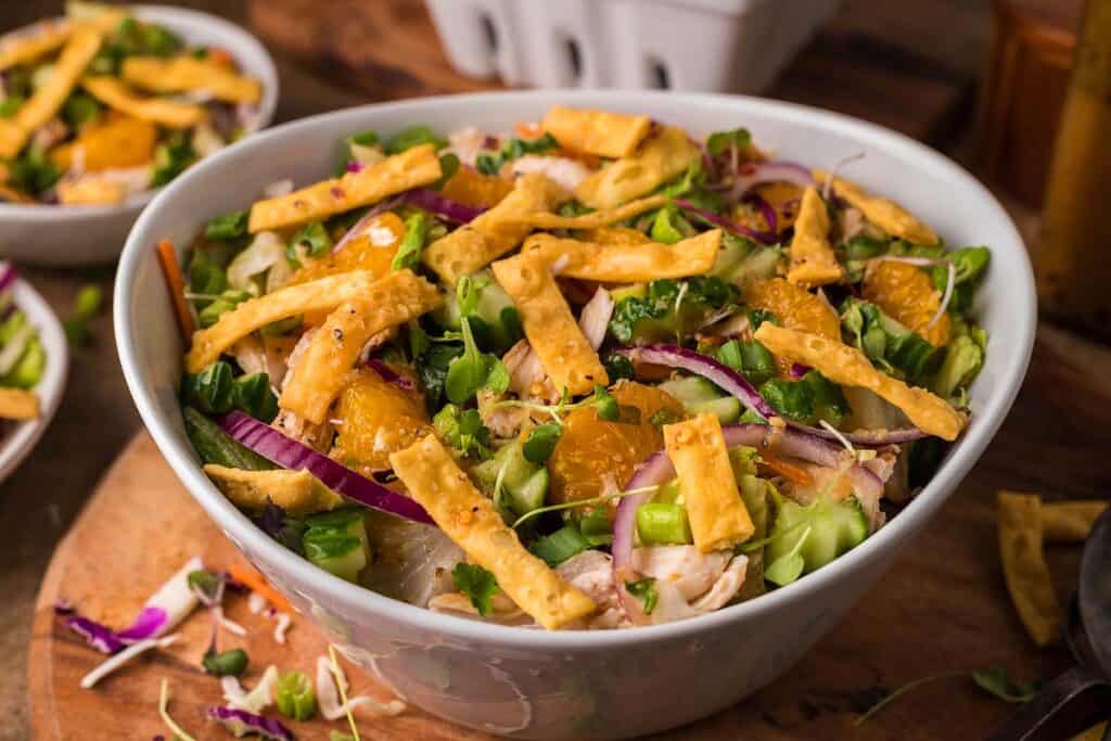 A bowl of salad with mixed greens, sliced red onions, mandarin orange segments, chopped green onions, and crispy wonton strips, topped with sesame seeds&mdash;a fresh take on Asian-inspired dishes.