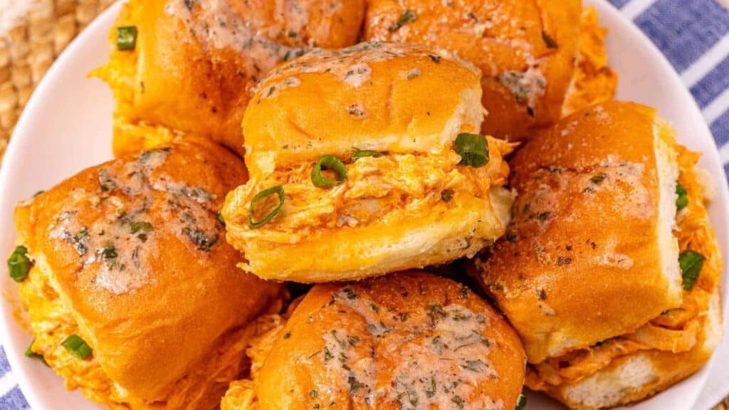 A plate of slider sandwiches filled with shredded buffalo chicken and topped with melted butter and herbs, garnished with chopped green onions.