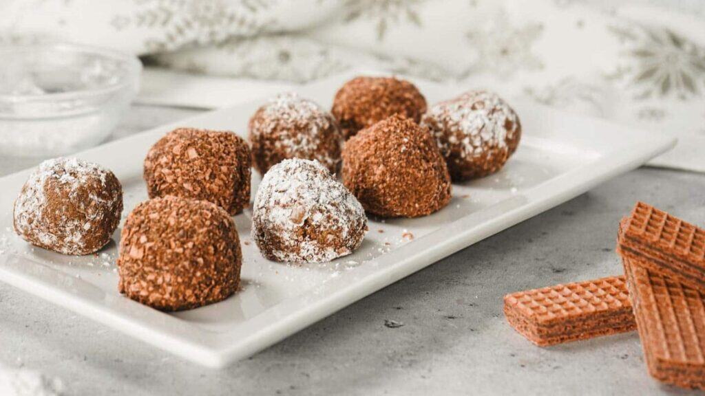 A rectangular white plate with several chocolate truffles, some dusted with powdered sugar, next to two chocolate wafer biscuits on a light gray surface.
