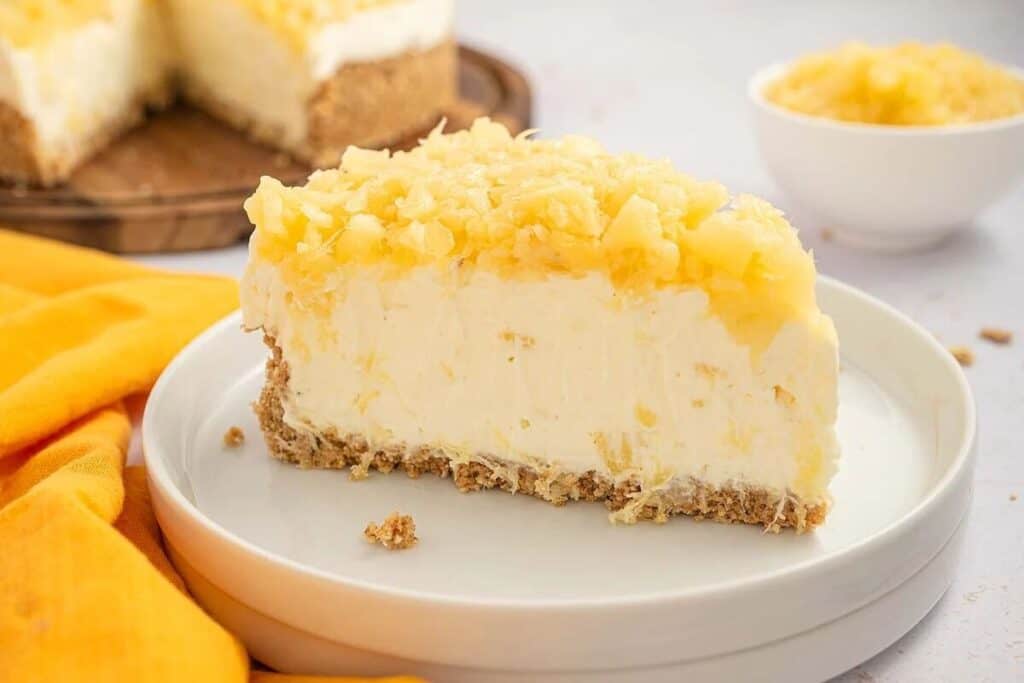 A slice of Simple Cheesecake with pineapple and a crumbly crust sits on a white plate, accompanied by a bowl of pineapple and the rest of the cheesecake in the background.
