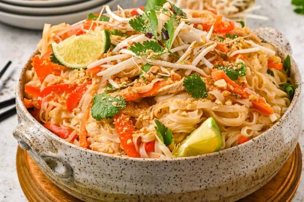 A bowl of pad Thai noodles garnished with lime wedges, bean sprouts, cilantro, peanuts, and shredded vegetables sits on a wooden tray&mdash;perfect for those who love Asian-inspired dishes.