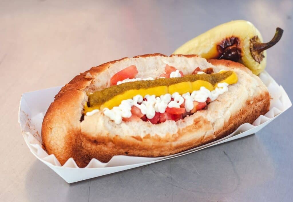 A hot dog topped with diced tomatoes, mayonnaise, mustard, and relish, served in a paper tray with a roasted pepper on the side.