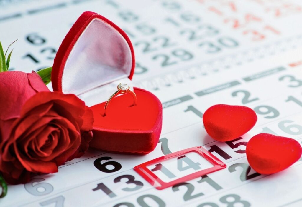 A gold ring in a red heart-shaped box, a red rose, two small red hearts, and a marked February 14 date on a calendar.
