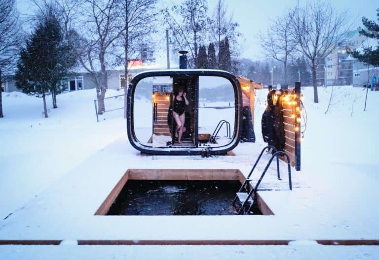 Embracing sauna culture, a person in swimwear stands at the entrance of a modern sauna beside an icy outdoor plunge pool surrounded by snow on a winter day.