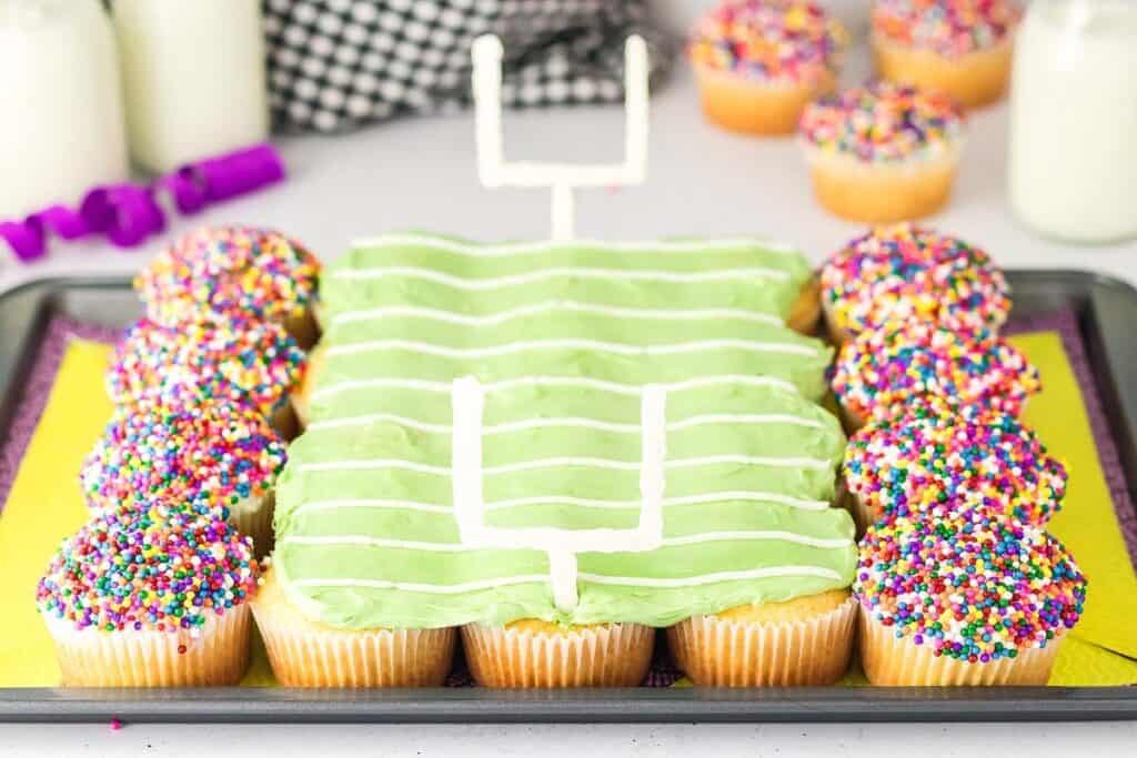 Cupcakes arranged and decorated to resemble a football field, with green icing, white goalposts, and multicolored sprinkle cupcakes on the sides.