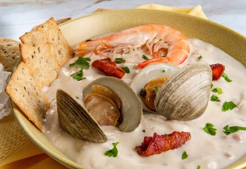 Celebrate National Clam Chowder Day with a bowl of creamy seafood chowder topped with two clams, a shrimp, pieces of bacon, fresh parsley, and served with crackers on the side.