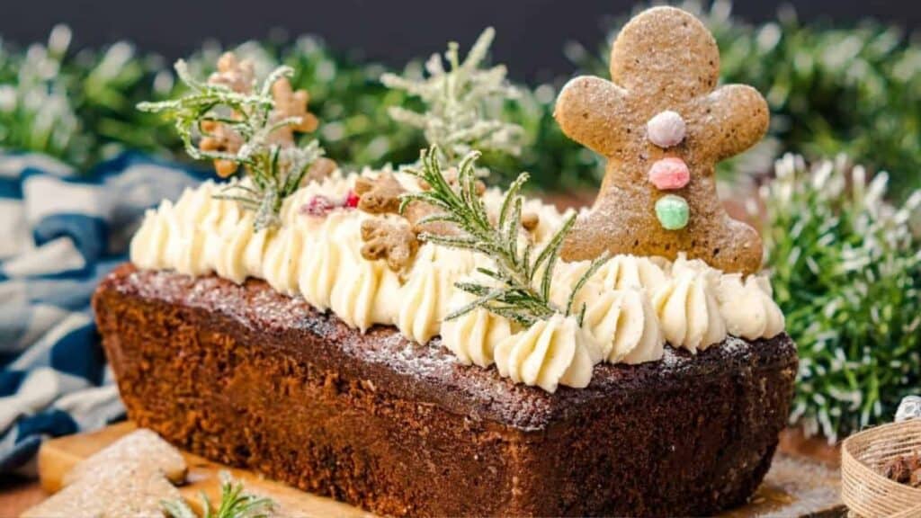 A rectangular loaf cake topped with piped frosting, a gingerbread man cookie, small cookies, and sprigs of rosemary, set against a festive background.