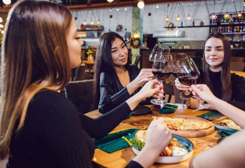 Four women celebrate Galentine's Day at a restaurant, clinking glasses of red wine over plates of pizza and salad.
