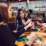 Four women celebrate Galentine's Day at a restaurant, clinking glasses of red wine over plates of pizza and salad.