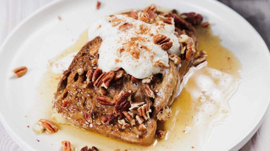 A slice of French toast topped with whipped cream, chopped pecans, a sprinkle of cinnamon, and syrup on a white plate.