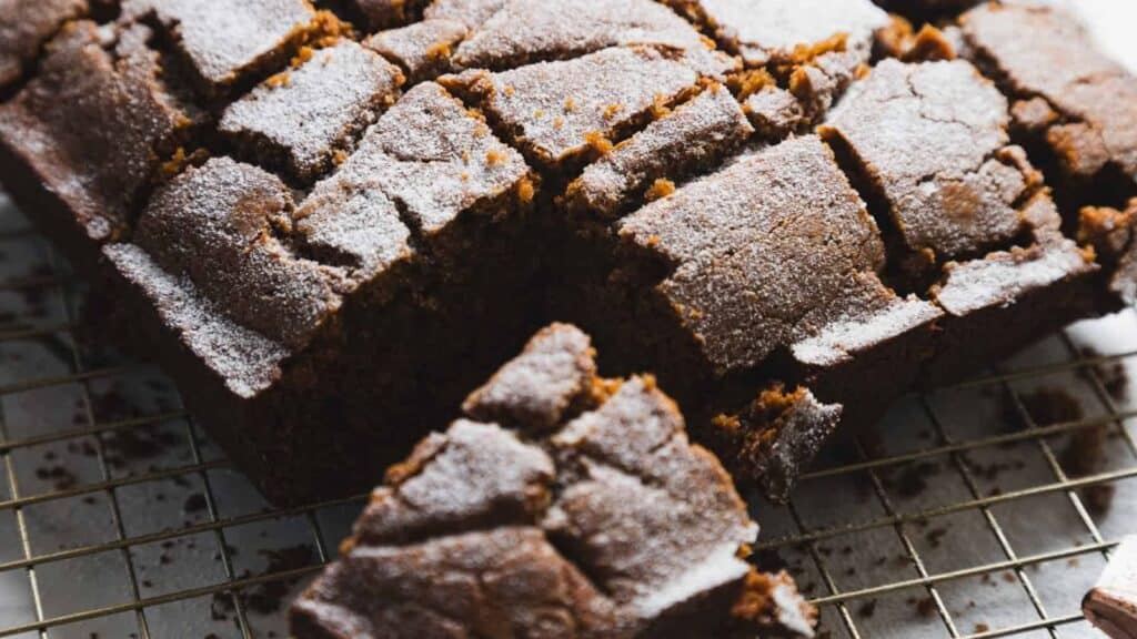 A loaf of sliced gingerbread cake with a cracked top and a dusting of powdered sugar, resting on a wire cooling rack.