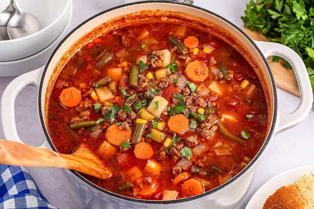 A large pot of vegetable beef soup with carrots, green beans, corn, potatoes, tomatoes, and ground beef, being stirred with a wooden spoon. Fresh parsley and bread are nearby.