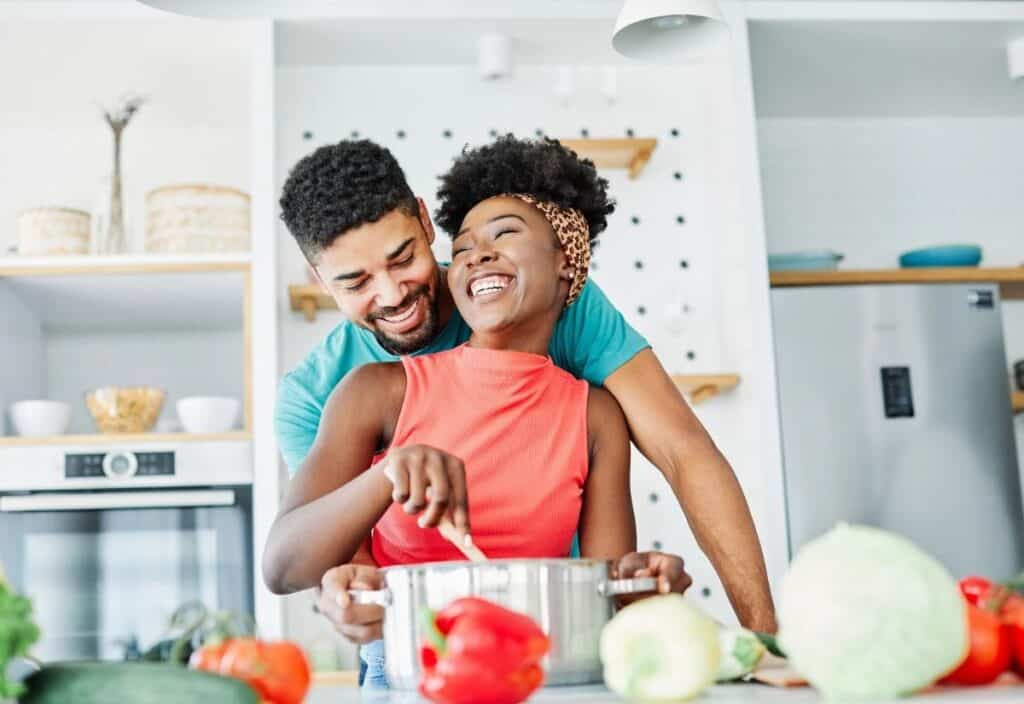 A man and woman stand in a modern kitchen, smiling as the woman stirs a pot on the counter surrounded by various fresh vegetables.