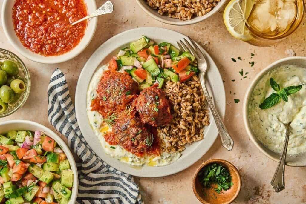 A plate with meatballs in tomato sauce, farro, and chopped cucumber salad, surrounded by bowls of tzatziki, olives, salad, tomato sauce, and a glass of lemon water&mdash;perfect for fans of tasty beef recipes.