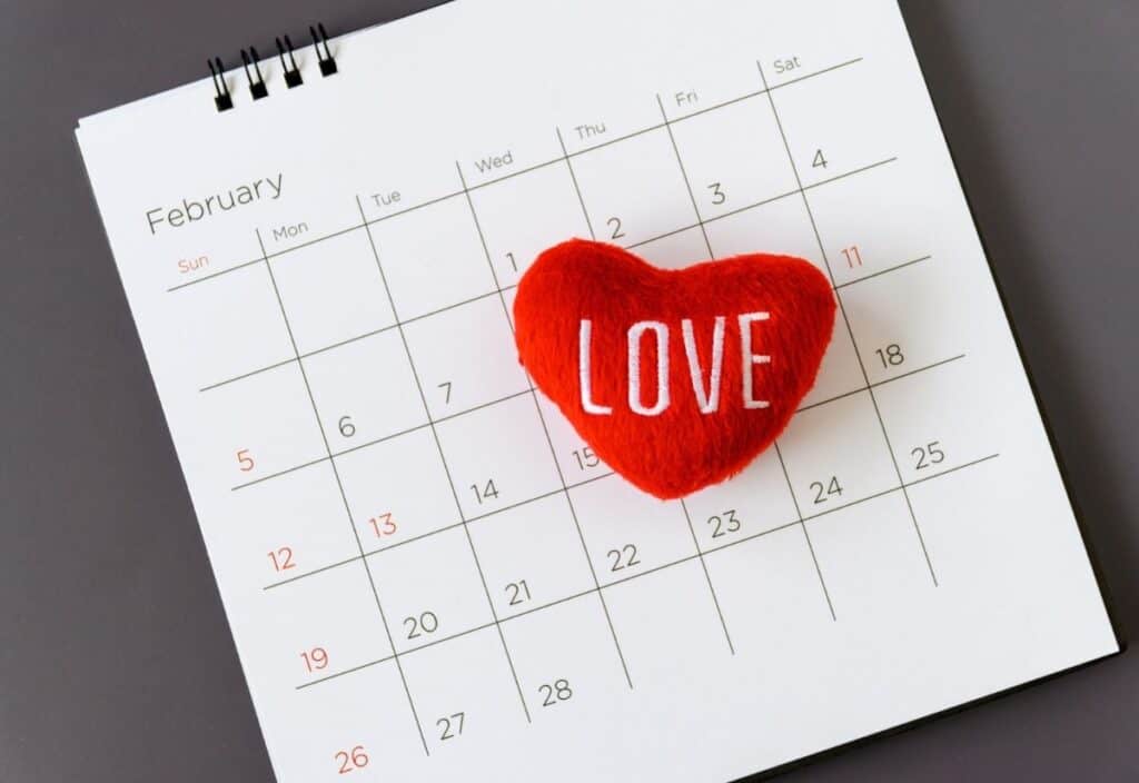 A red heart-shaped cushion with the word "LOVE" lies on a calendar opened to February, with Valentine's Day marked on the 14th.