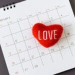 A red heart-shaped cushion with the word "LOVE" lies on a calendar opened to February, with Valentine's Day marked on the 14th.
