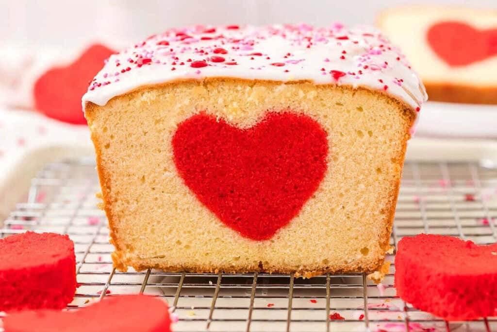 A loaf cake with white icing and red sprinkles features a red heart shape in the center, making it perfect for Valentines Treats. Shown on a cooling rack with heart-shaped pieces nearby.