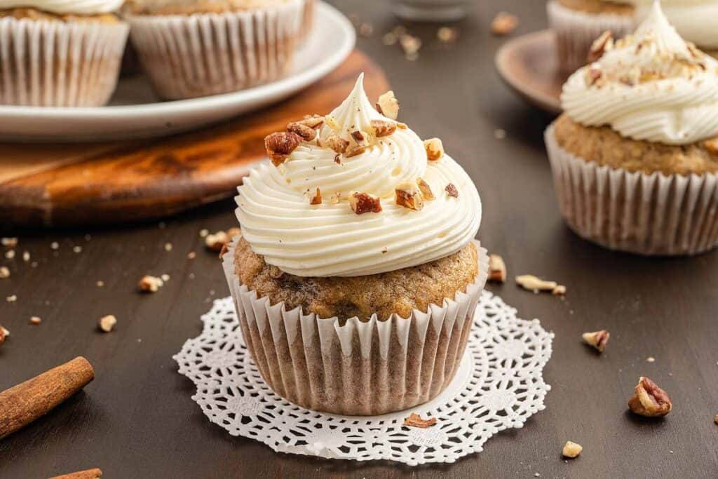 A cupcake with swirled white frosting and chopped nuts on top sits on a lace doily, with more cupcakes on plates in the background.