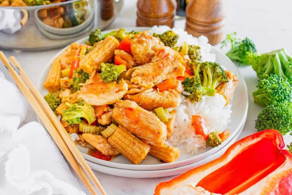A plate of white rice topped with stir-fried chicken, broccoli, baby corn, celery, and red bell pepper&mdash;an inviting example of Asian-inspired dishes&mdash;served with chopsticks and fresh vegetables nearby.