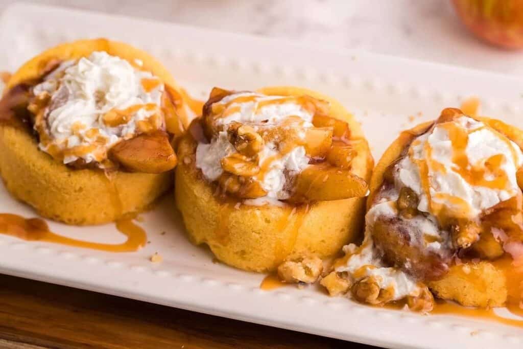 Three sponge cake cups filled with apple slices, walnuts, and whipped cream, drizzled with caramel sauce, arranged on a white rectangular plate&mdash;perfect alongside your favorite instant pot dishes.