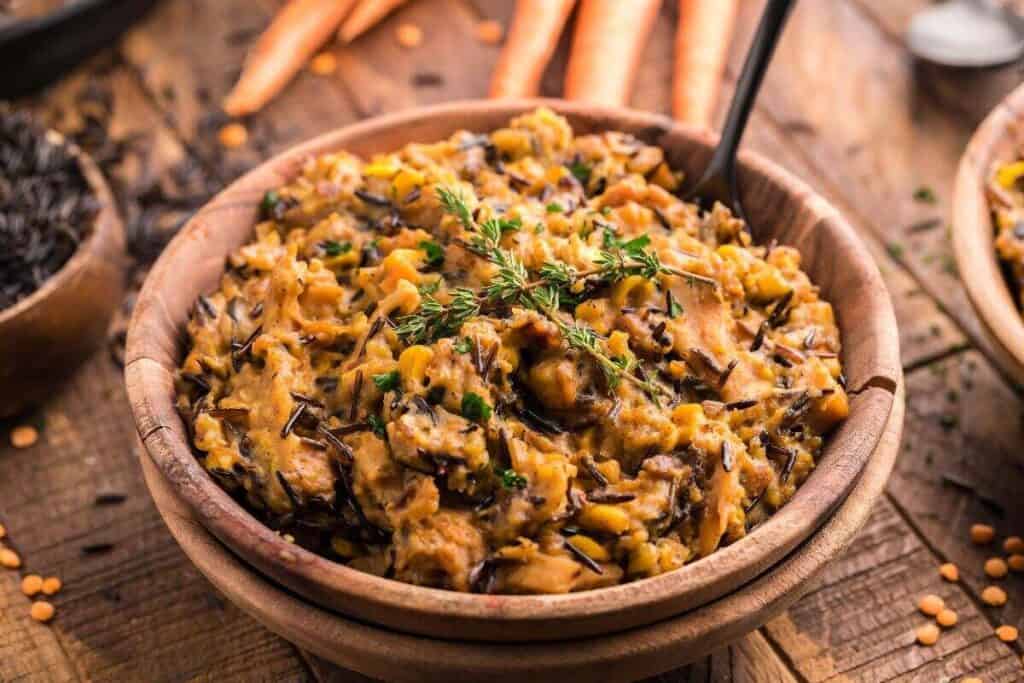 A wooden bowl filled with creamy wild rice and vegetable casserole, one of the coziest instant pot dishes, topped with fresh herbs, with a spoon inside and carrots in the background.